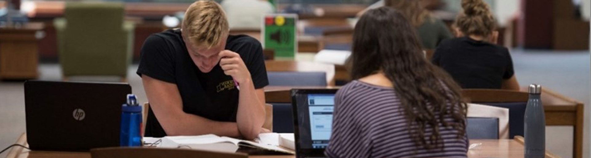 Two students, one male and one female, sit diagonally across from each other at a table, studying in the Centennial Library.